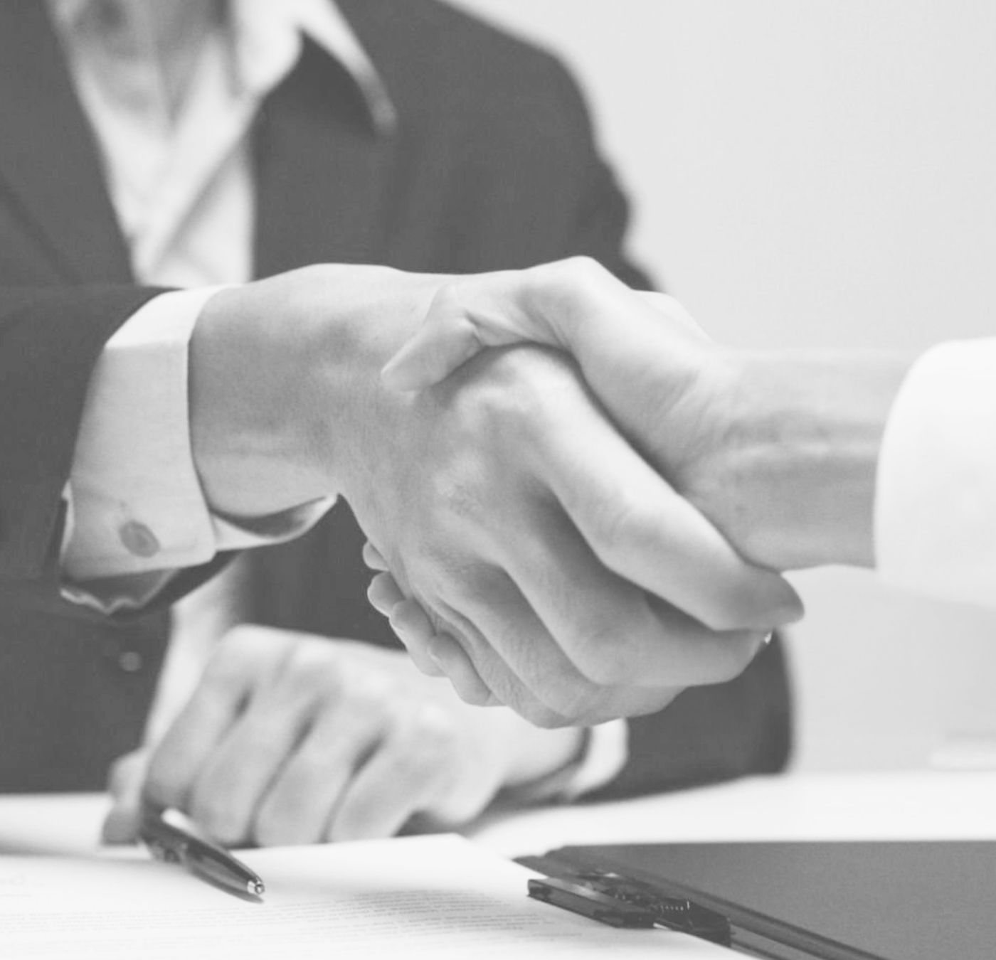 Two people in suits shaking hands across a desk with a pen and folder in view