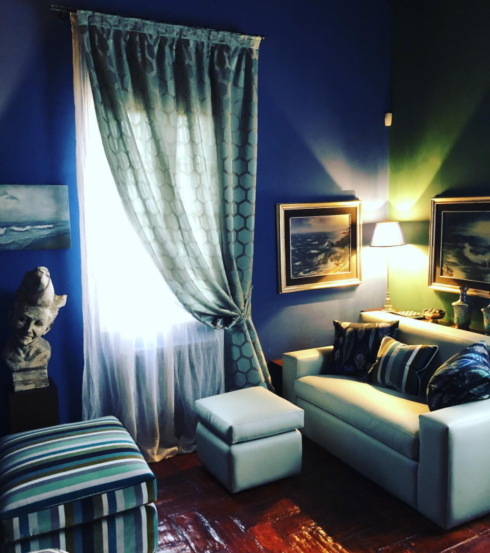 Soggiorno con pareti blu, divano bianco, tende a fantasia, opere d'arte e un pouf.