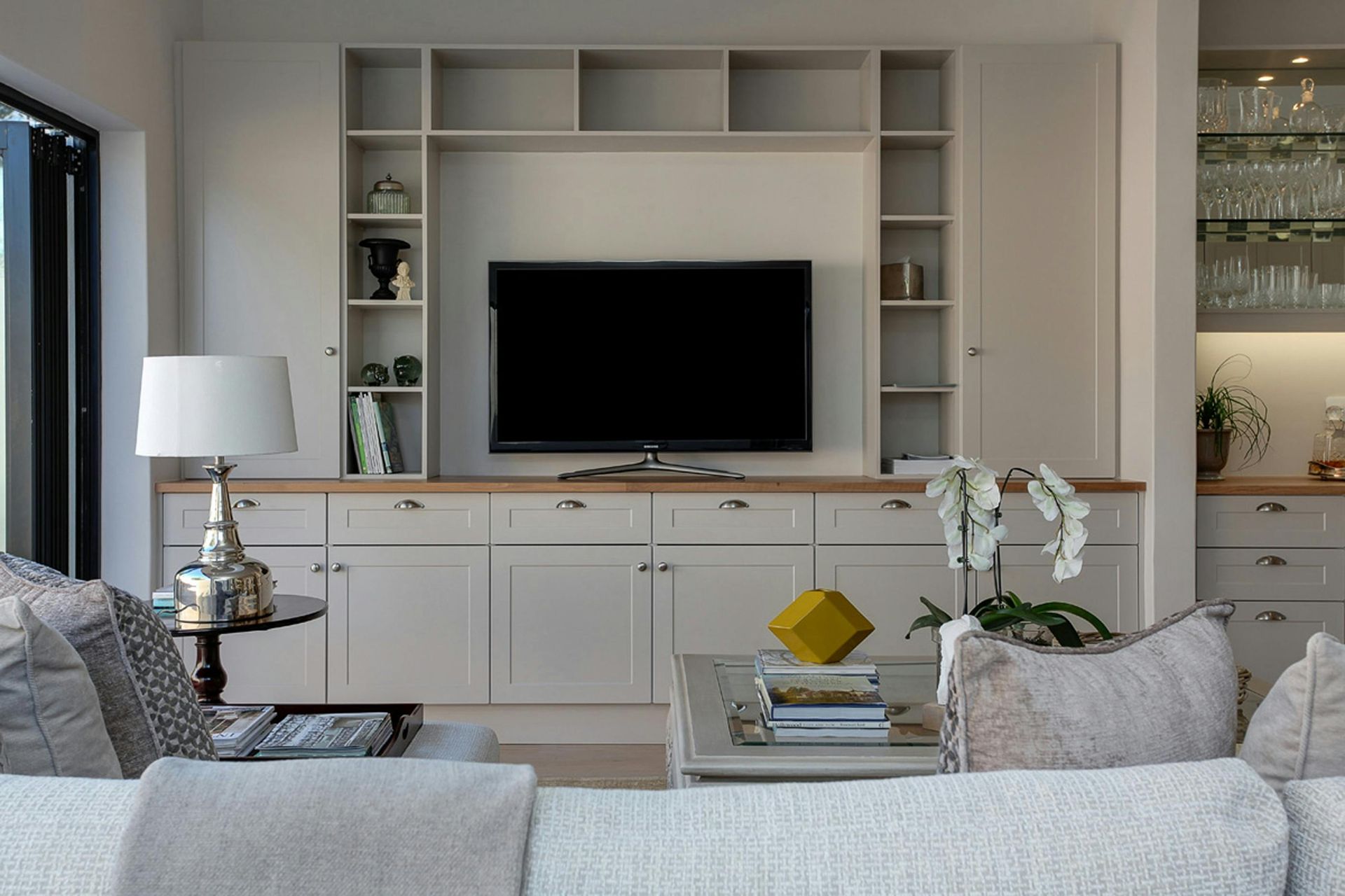Living room with built-in entertainment center, TV, shelves, and cabinets. Light colors, sofa, lamp, and coffee table.
