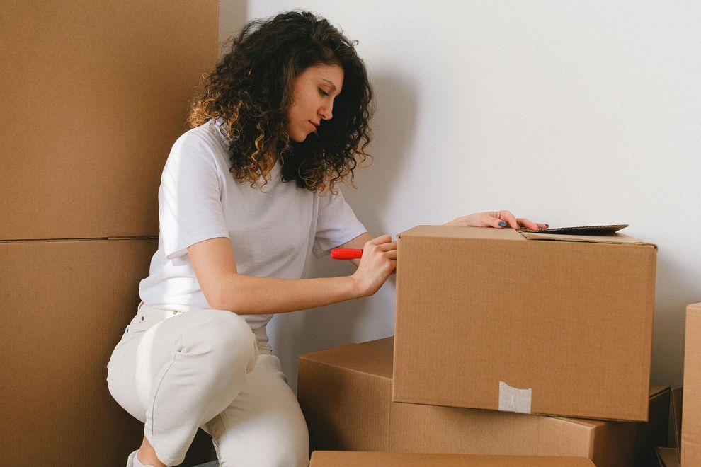 Woman kneeling, labeling cardboard box with marker near a stack of boxes against a white wall.