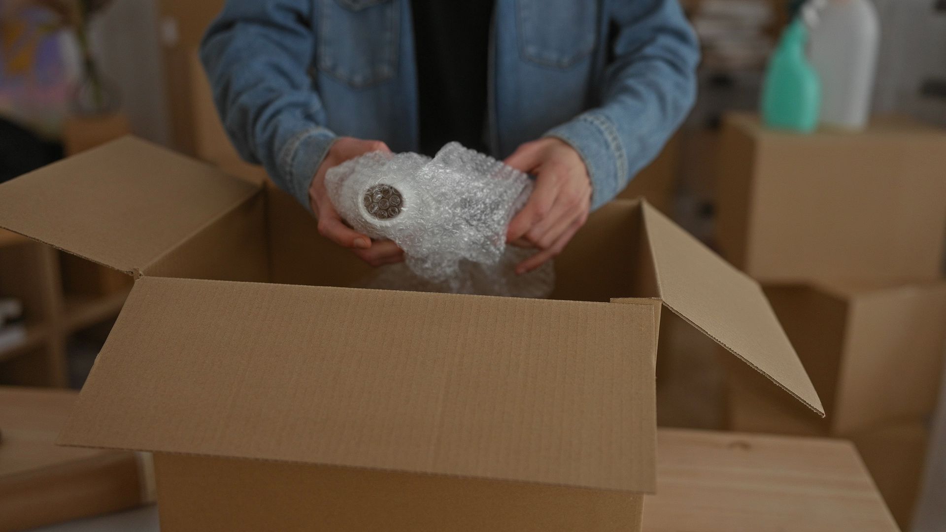 Person packing items in a cardboard box, wrapping them in bubble wrap, indoors.
