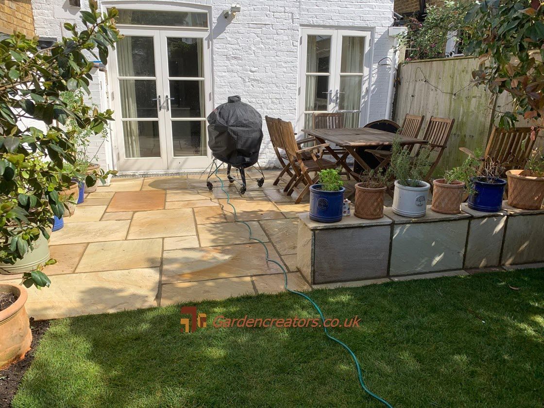 A stone patio with a grill, dining set, and potted plants outside white French doors, next to a grassy lawn.