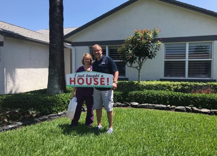 A man and woman are standing in front of a house holding a sign that says `` i just bought a house ''.