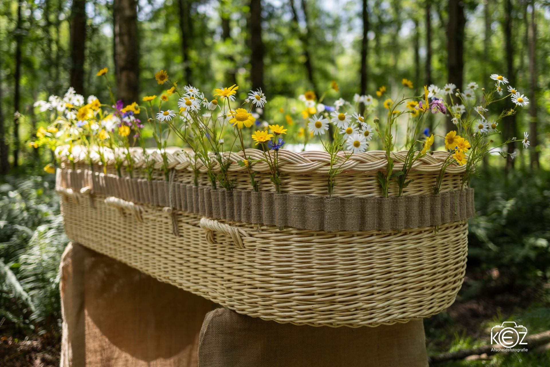 Een rieten doodskist, versierd met wilde bloemen en afgewerkt met jute, staat op met stof beklede standaards in een bosrijke omgeving.