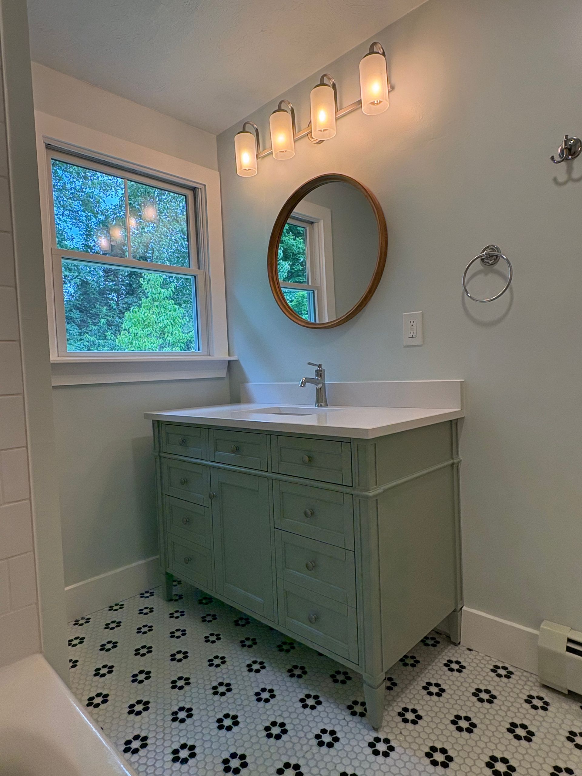 Bright bathroom with green vanity, round mirror, patterned tile floor, and window by a tree view