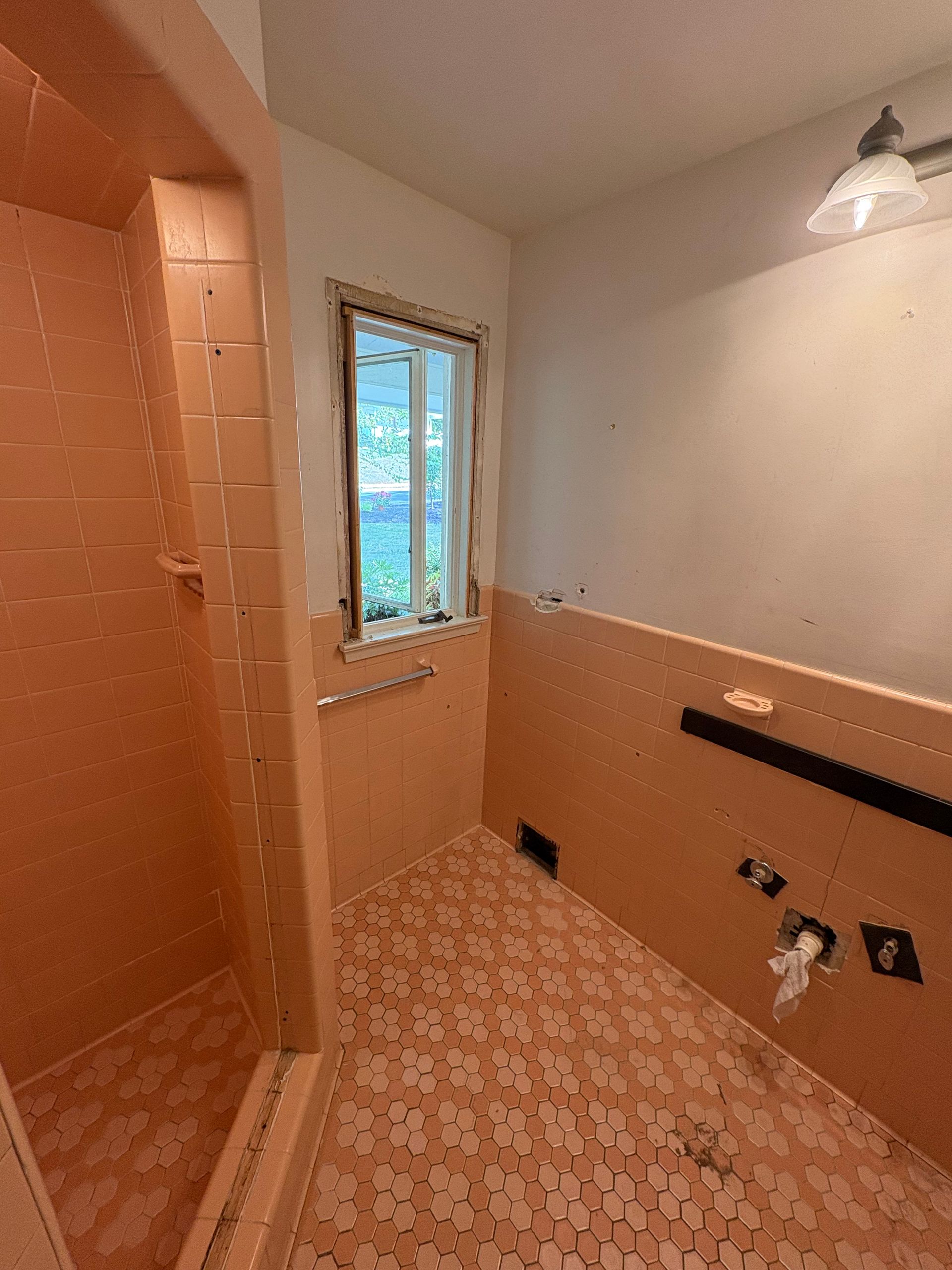 Narrow tiled bathroom under renovation with peach walls, window, sink cutout, and exposed plumbing fixtures
