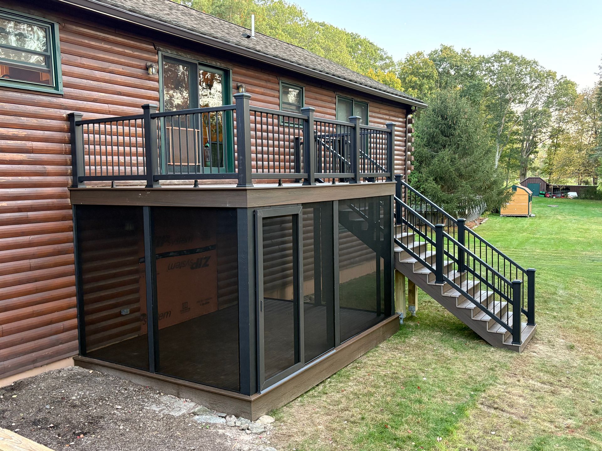 Back deck and exterior staircase on a log cabin, with large glass doors below and a grassy yard.