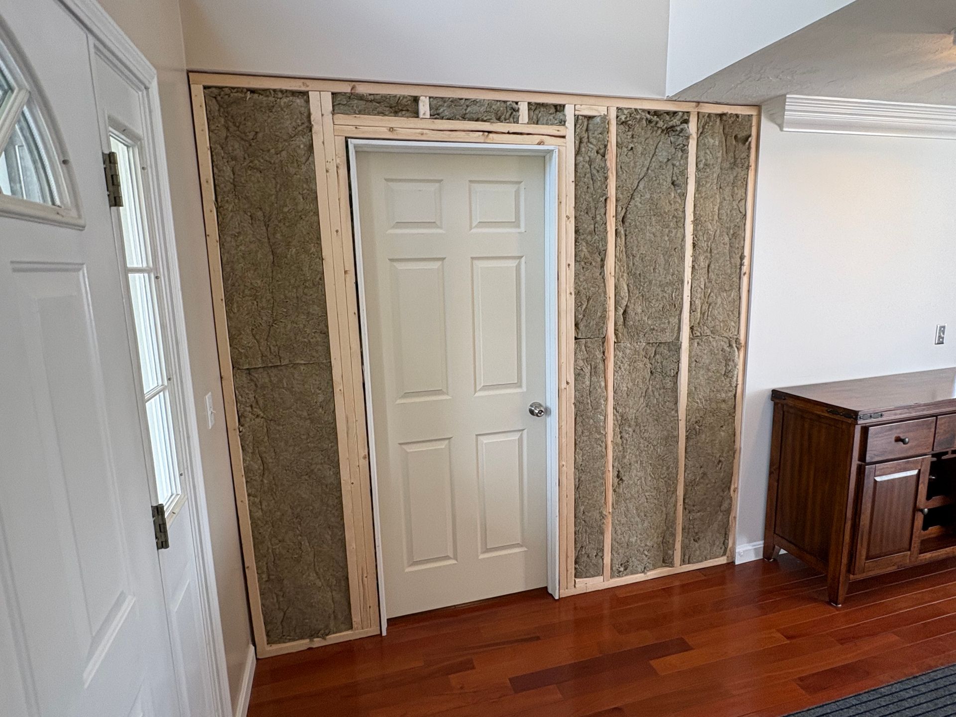 Living room wall with exposed studs framing a white door, beside a hardwood floor and cabinet.