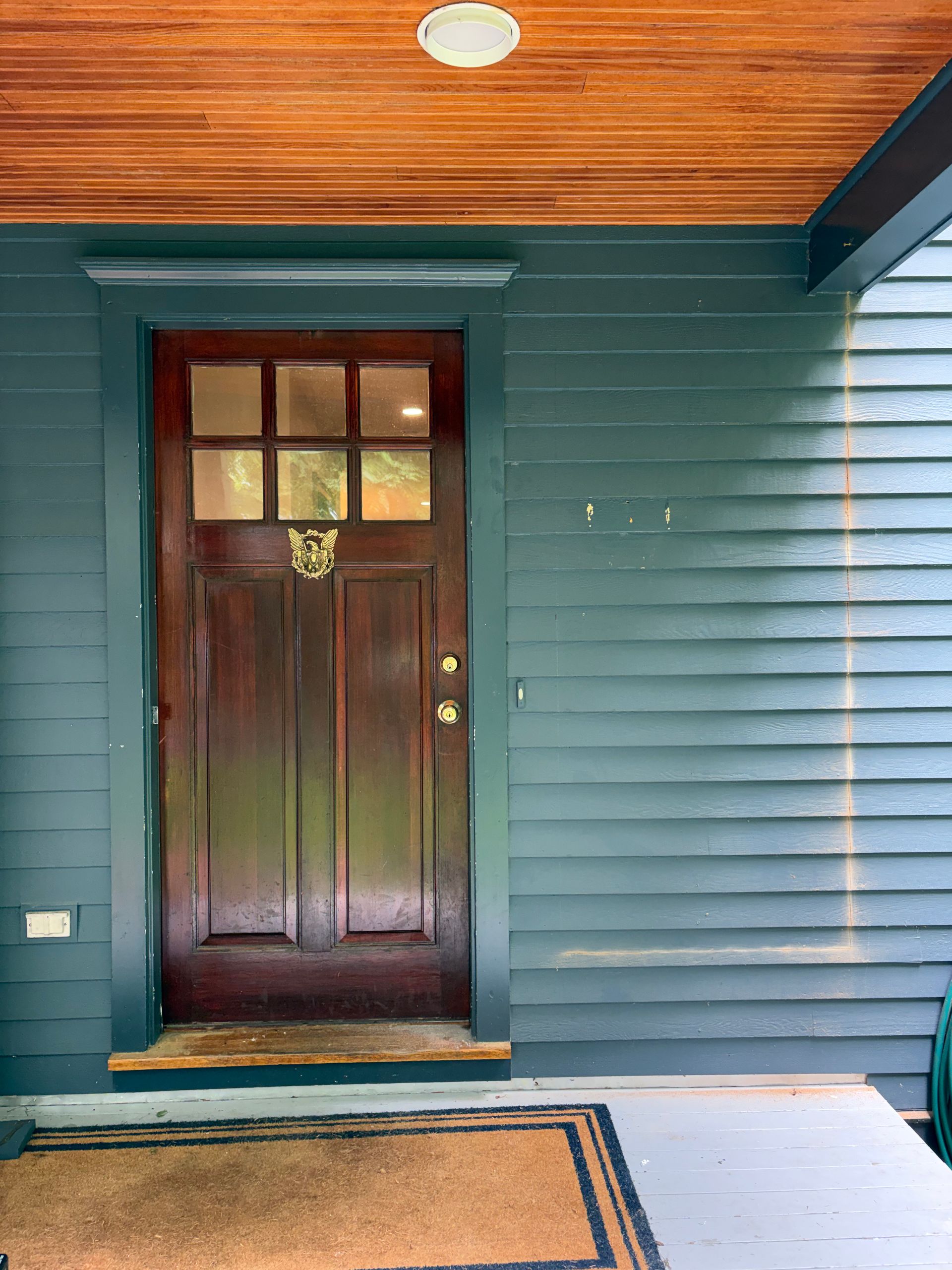 Front porch with a dark wooden front door, teal siding, and a tan doormat.