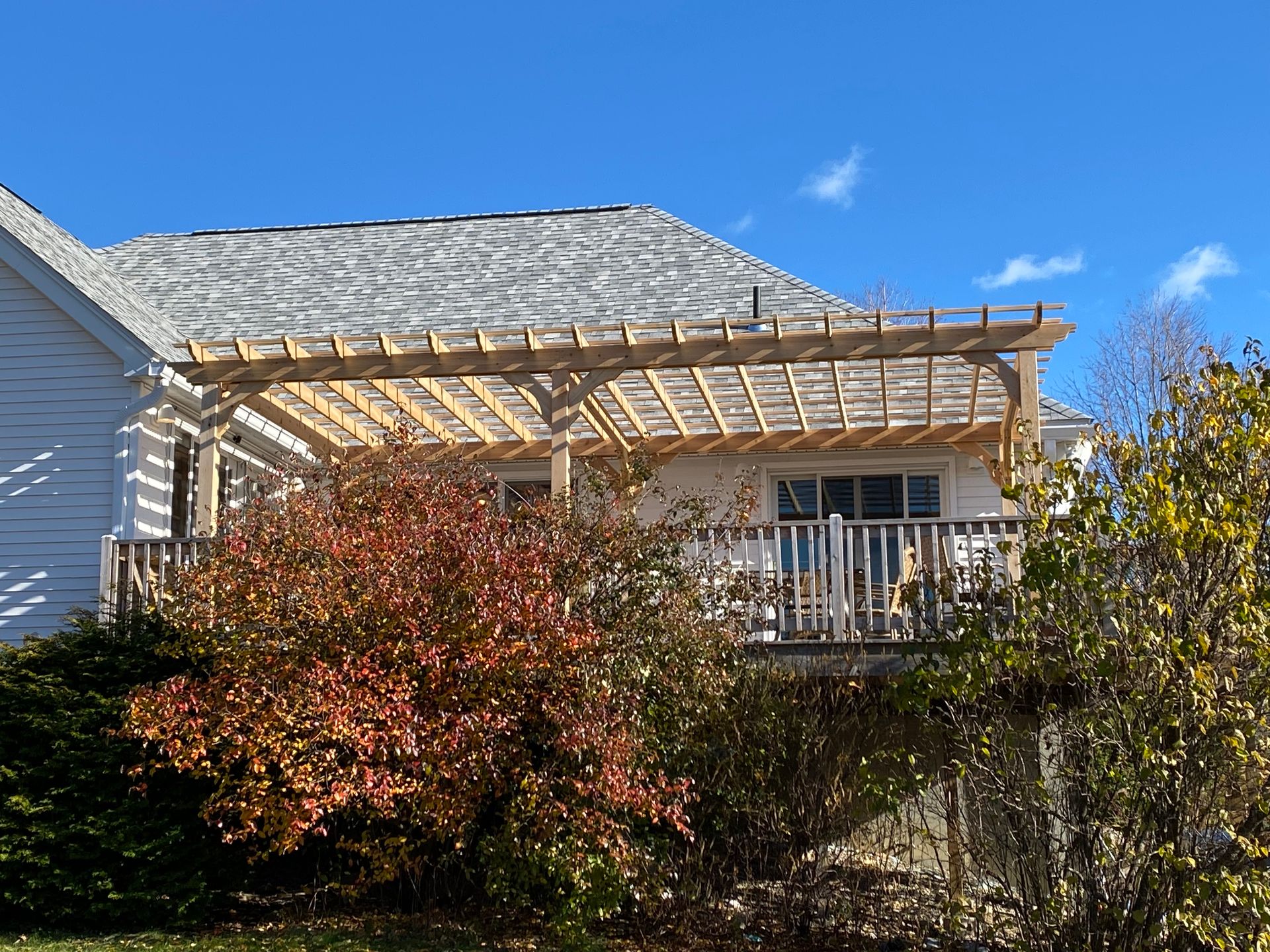 House with a wooden pergola over a back deck, framed by autumn shrubs under a blue sky.