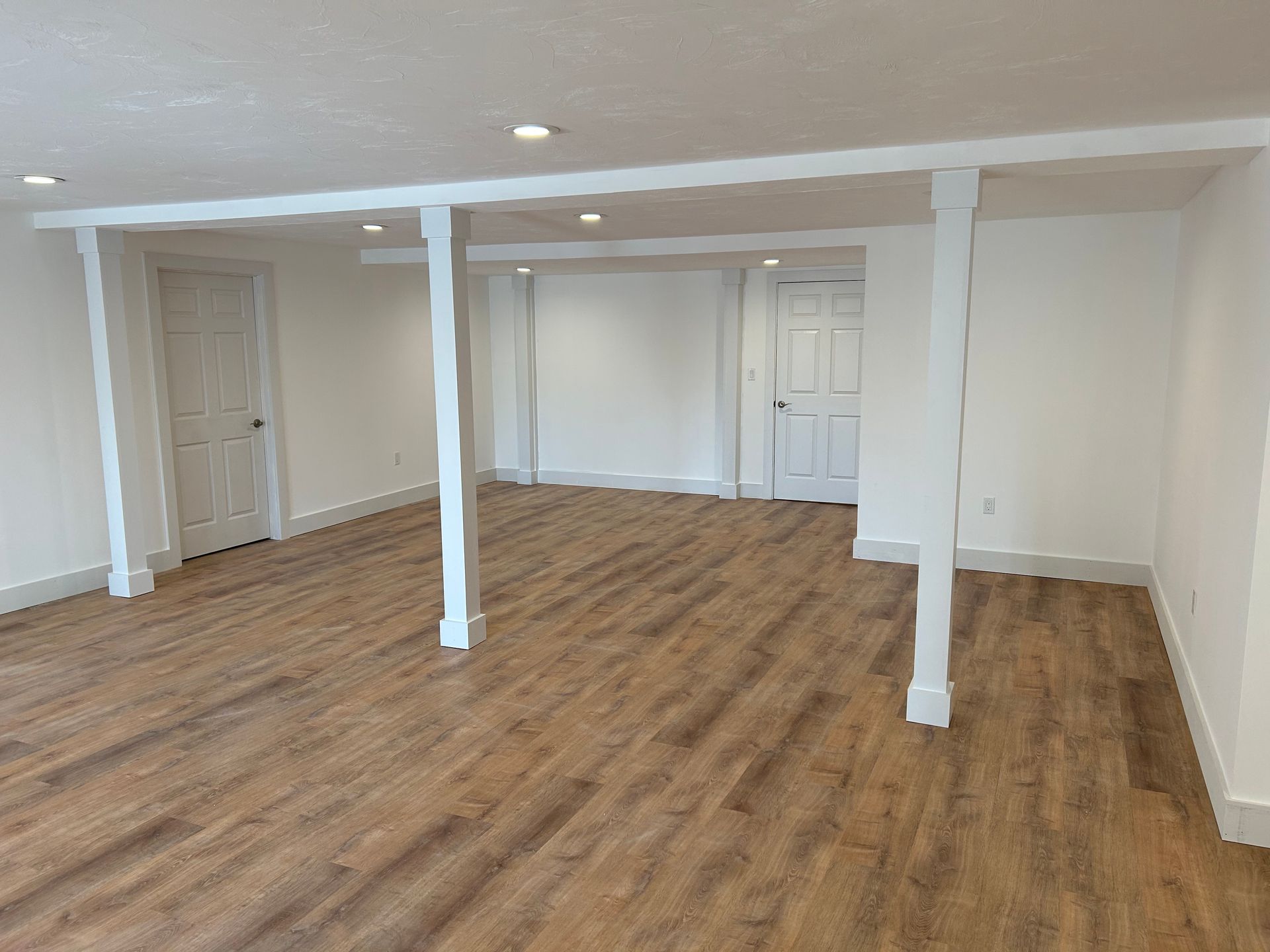 Empty basement room with wood flooring, white walls, support columns, and closed doors.