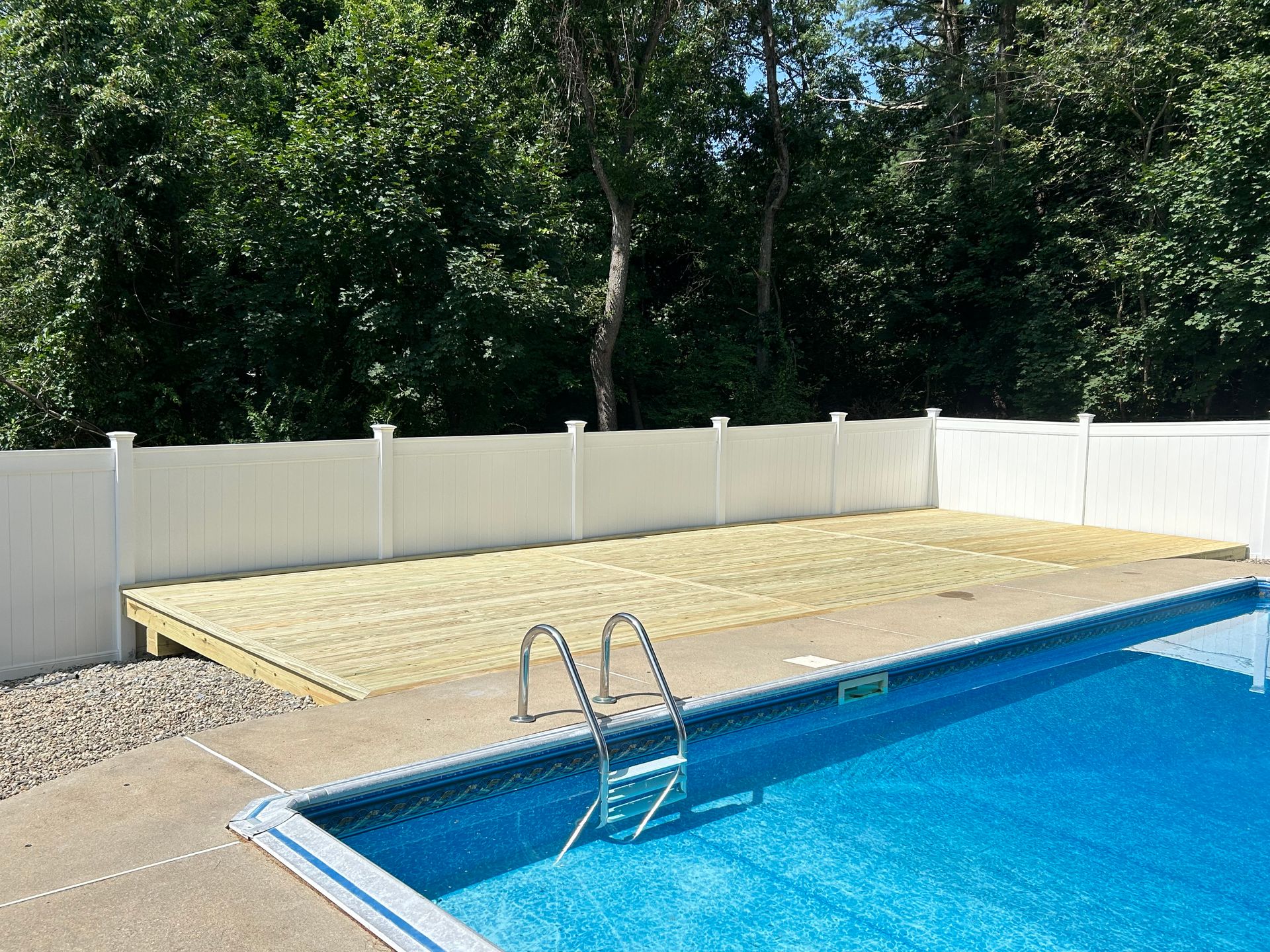 Backyard pool with blue water, metal ladder, and raised wooden deck beside a white fence and trees