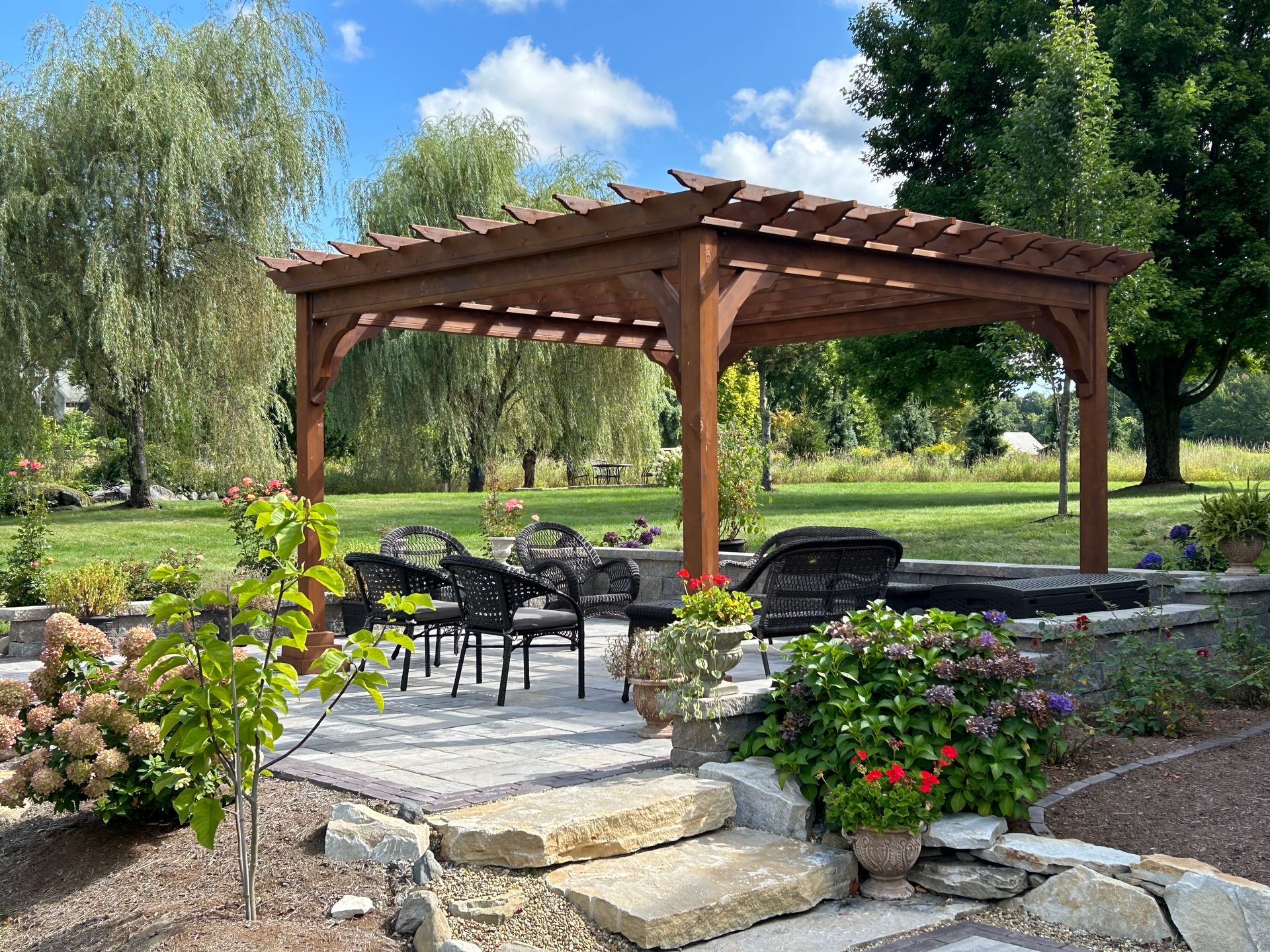 Wooden pergola in a landscaped garden with patio chairs, stone steps, and flowering shrubs under a blue sky