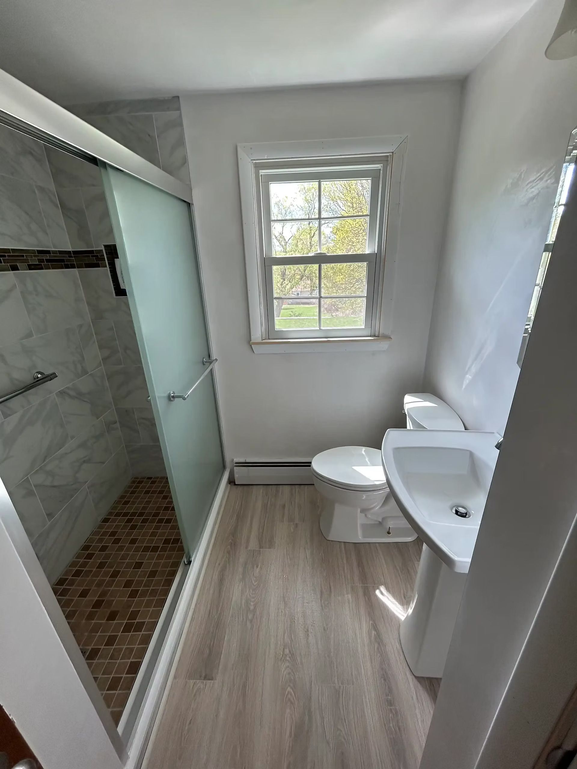 Narrow bathroom with walk-in shower, toilet, sink, and window, featuring white walls and wood-look flooring