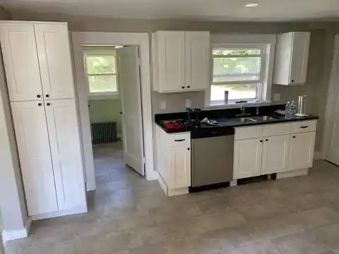 Bright kitchen with white cabinets, black countertop, stainless dishwasher, and open doorway to another room