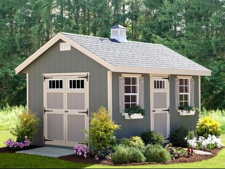 Small gray garden shed with white trim, double doors, windows, and flower beds in a wooded yard