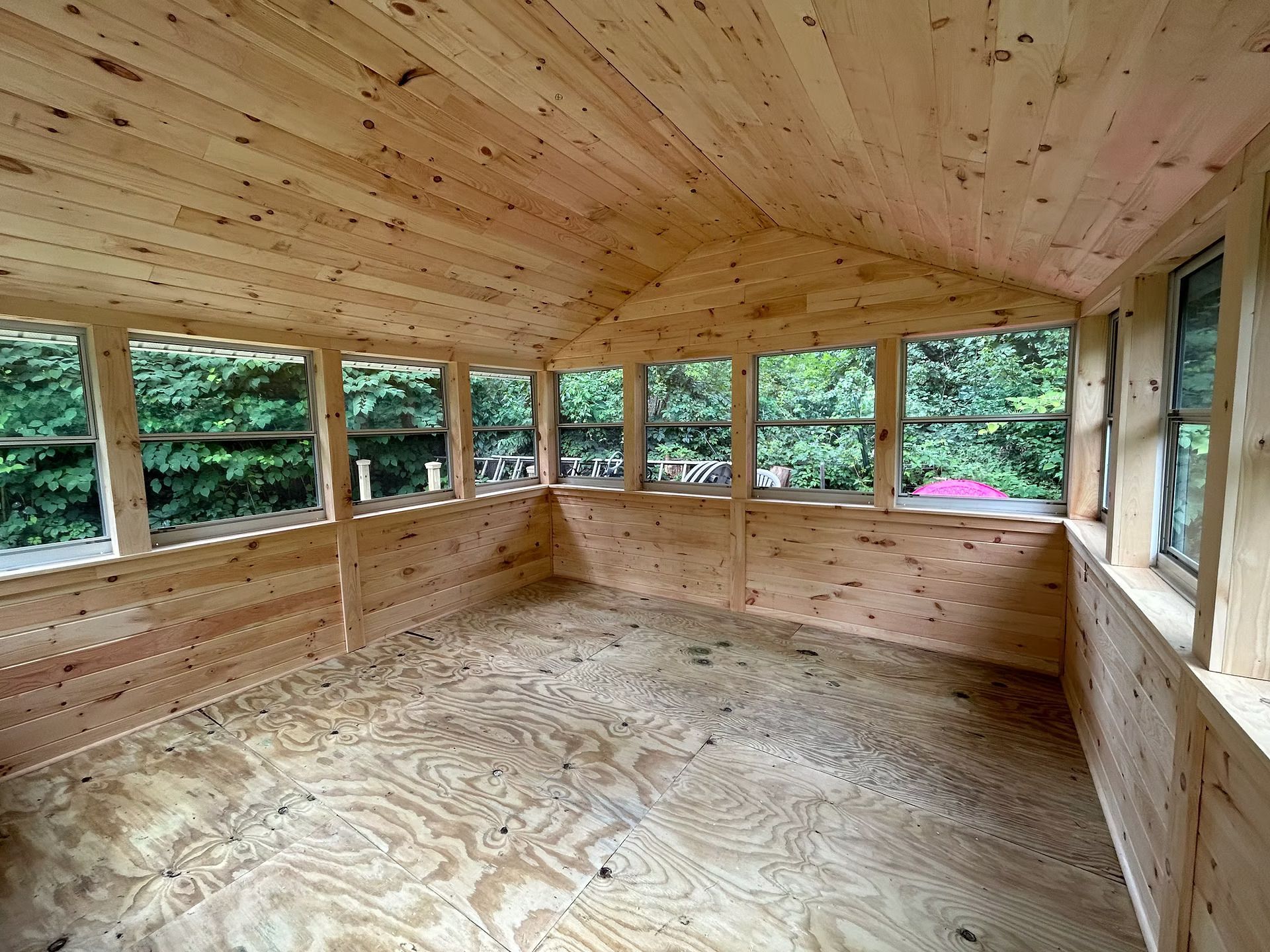 Empty unfinished wooden room with large windows overlooking trees and a plywood floor and ceiling