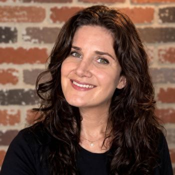 Portrait of a smiling woman with dark wavy hair in front of a brick wall