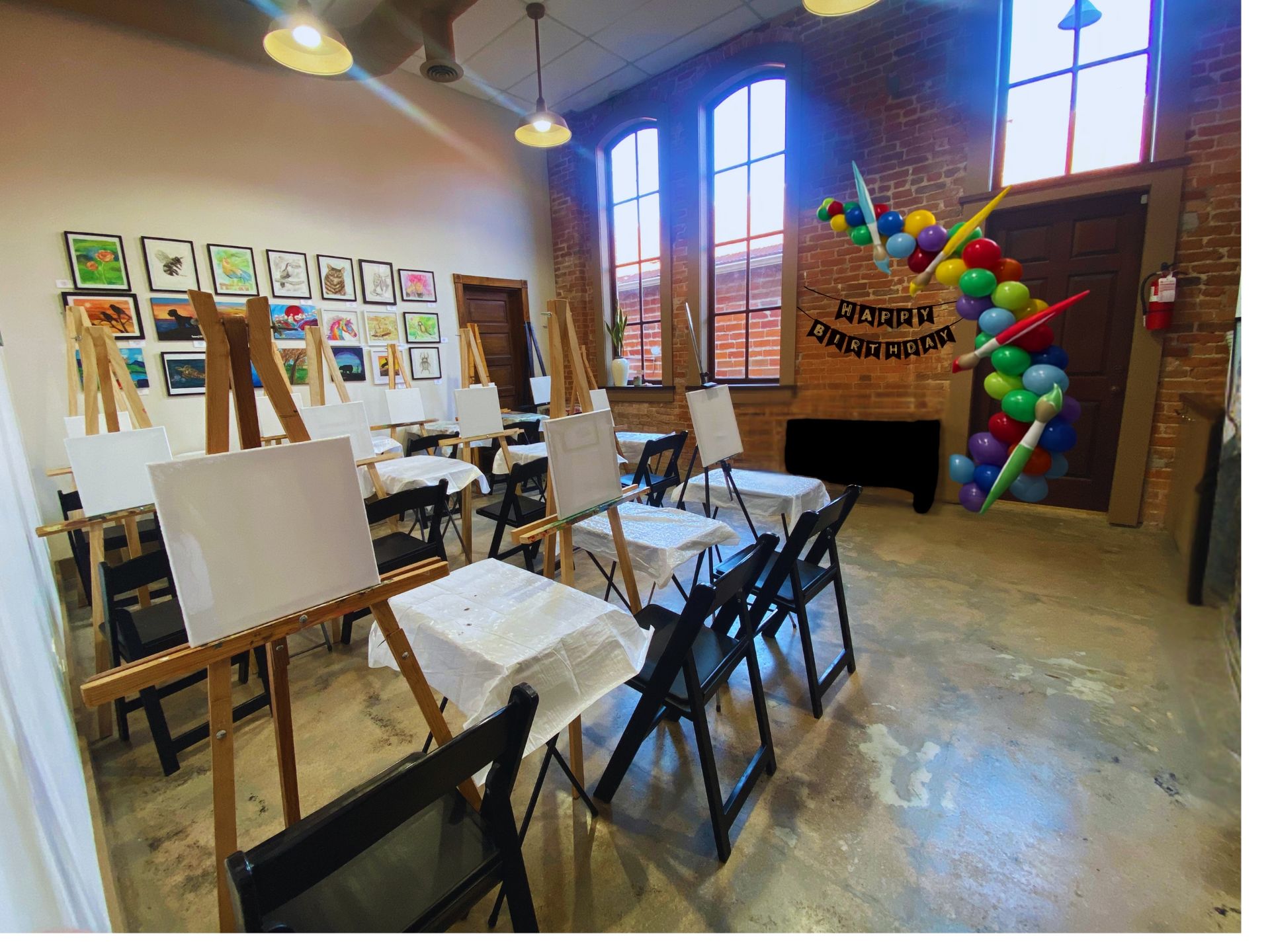 Art studio with easels and chairs, colorful balloon arch near a brick wall window
