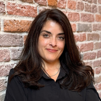 Portrait of a smiling woman with dark hair in a black shirt against a brick wall