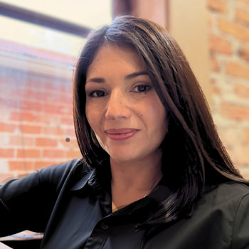 Woman with long dark hair in a black shirt, smiling indoors near a brick wall and window.