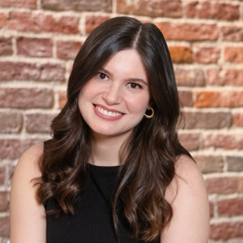 Smiling person with long dark hair in a black top, posed against a brick wall background.
