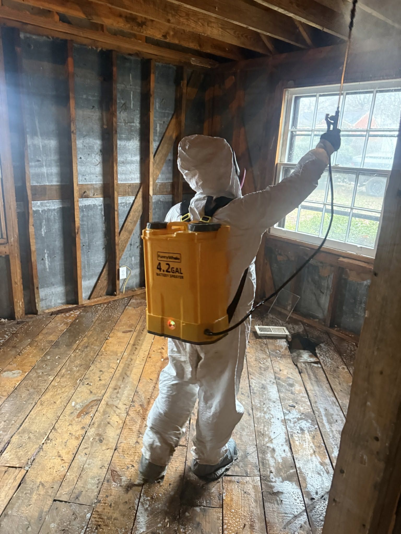 Person in protective suit spraying inside a stripped room with exposed studs and a window.