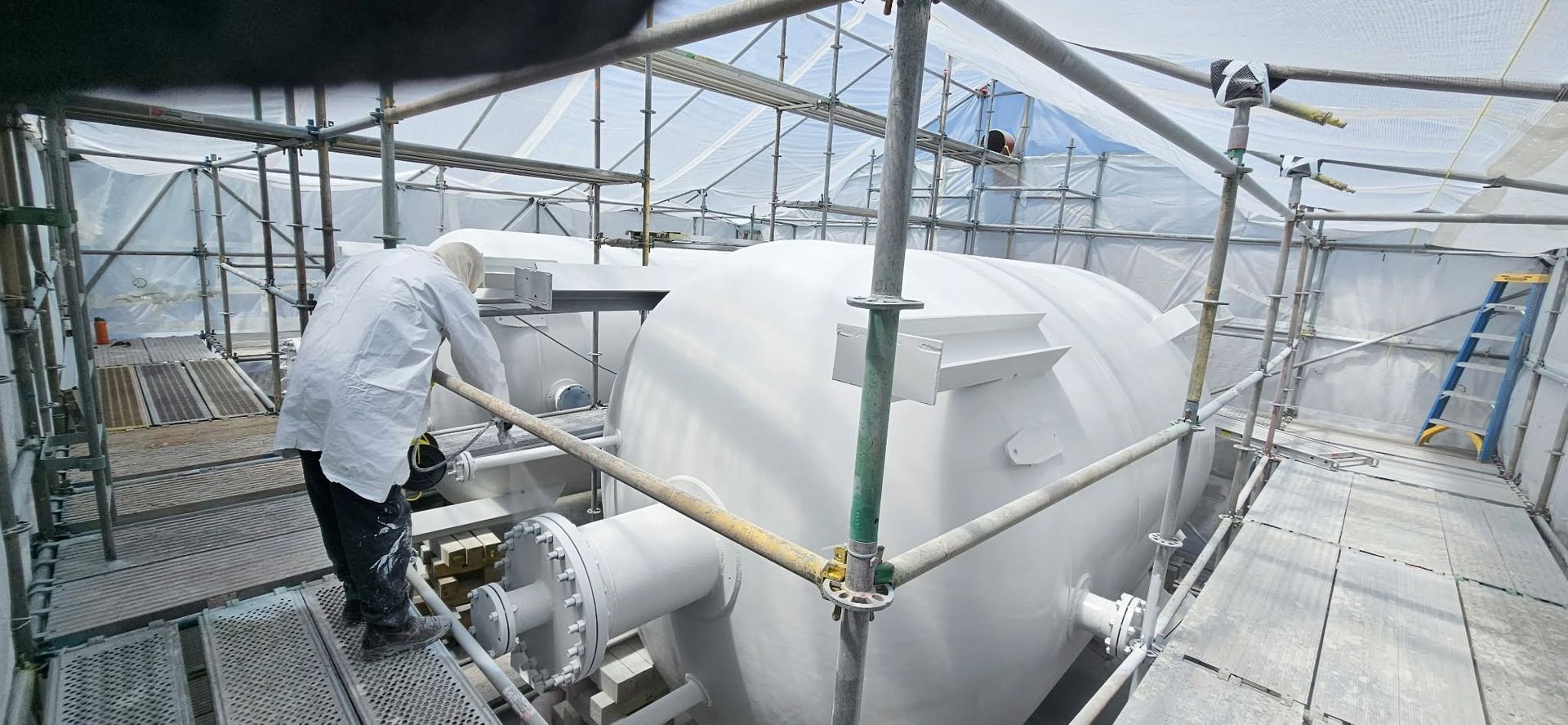 Worker in white protective gear inspecting equipment inside a greenhouse-like industrial structure.