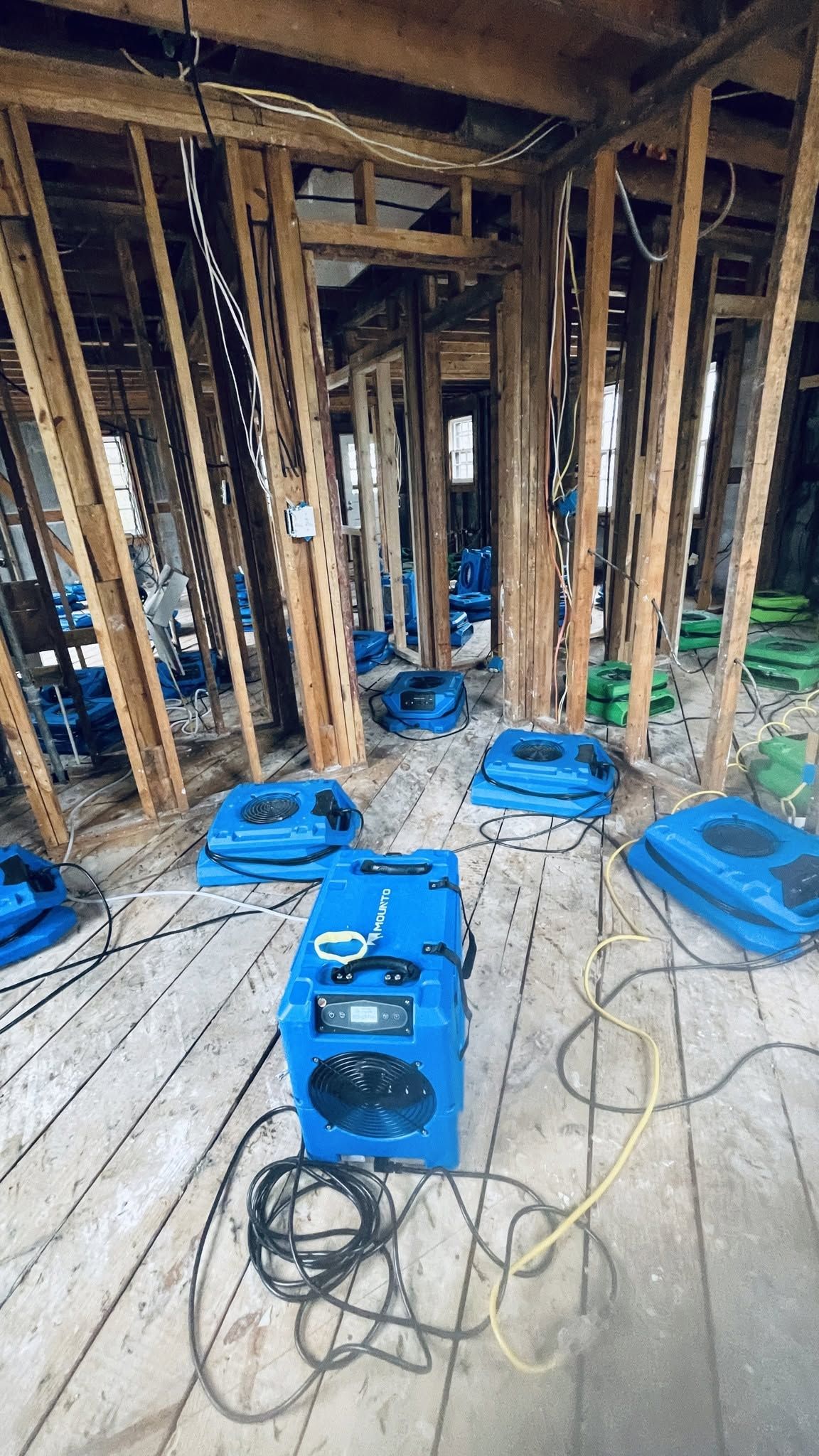 Damaged interior with exposed wood framing, blue equipment, and tangled cords on a debris-covered floor.