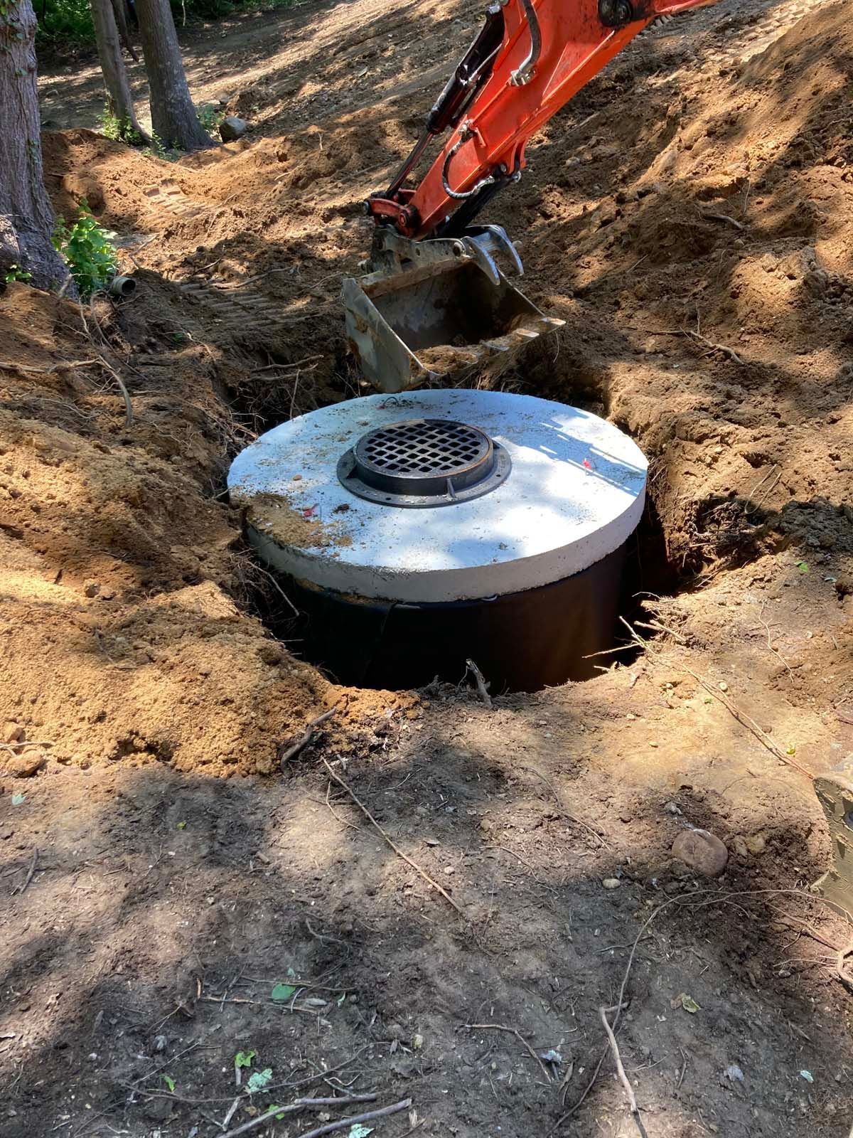 An excavator working to install a cylindrical structure with a grate in a dirt hole.