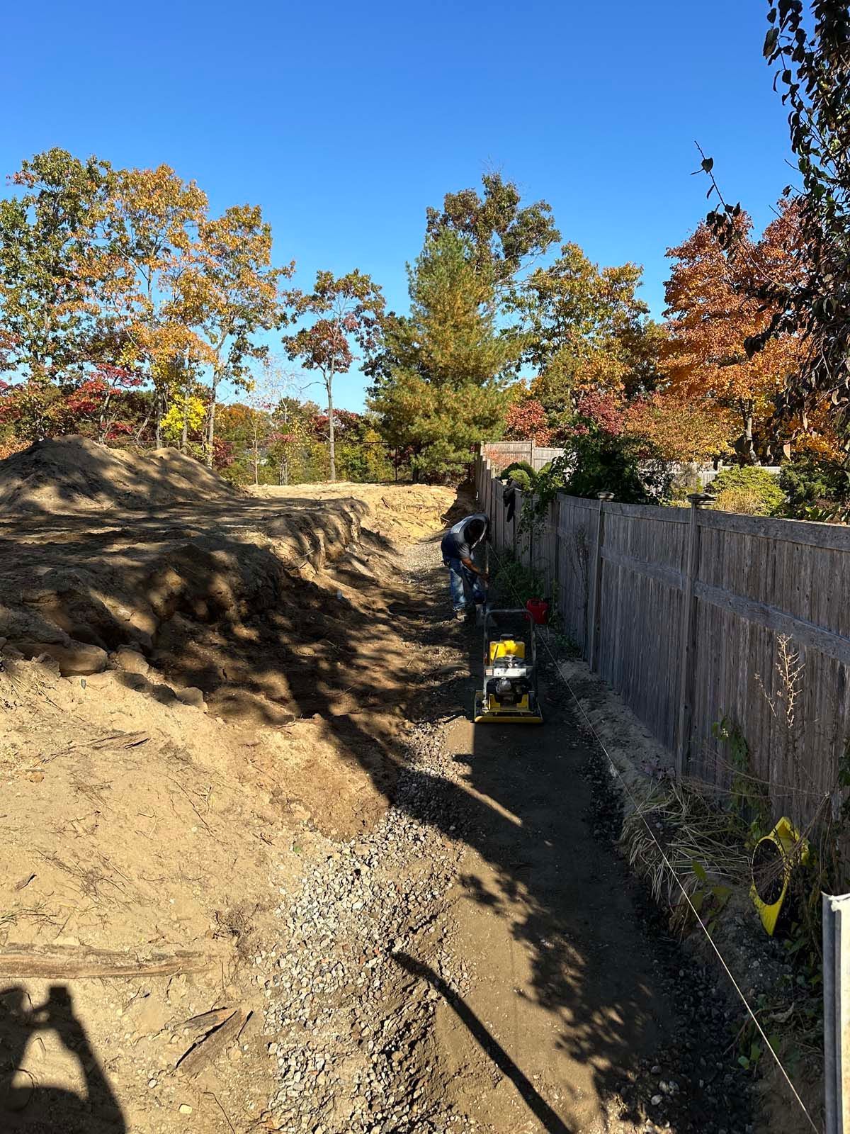 Construction site with person operating a compactor along a dirt trench next to a fence.