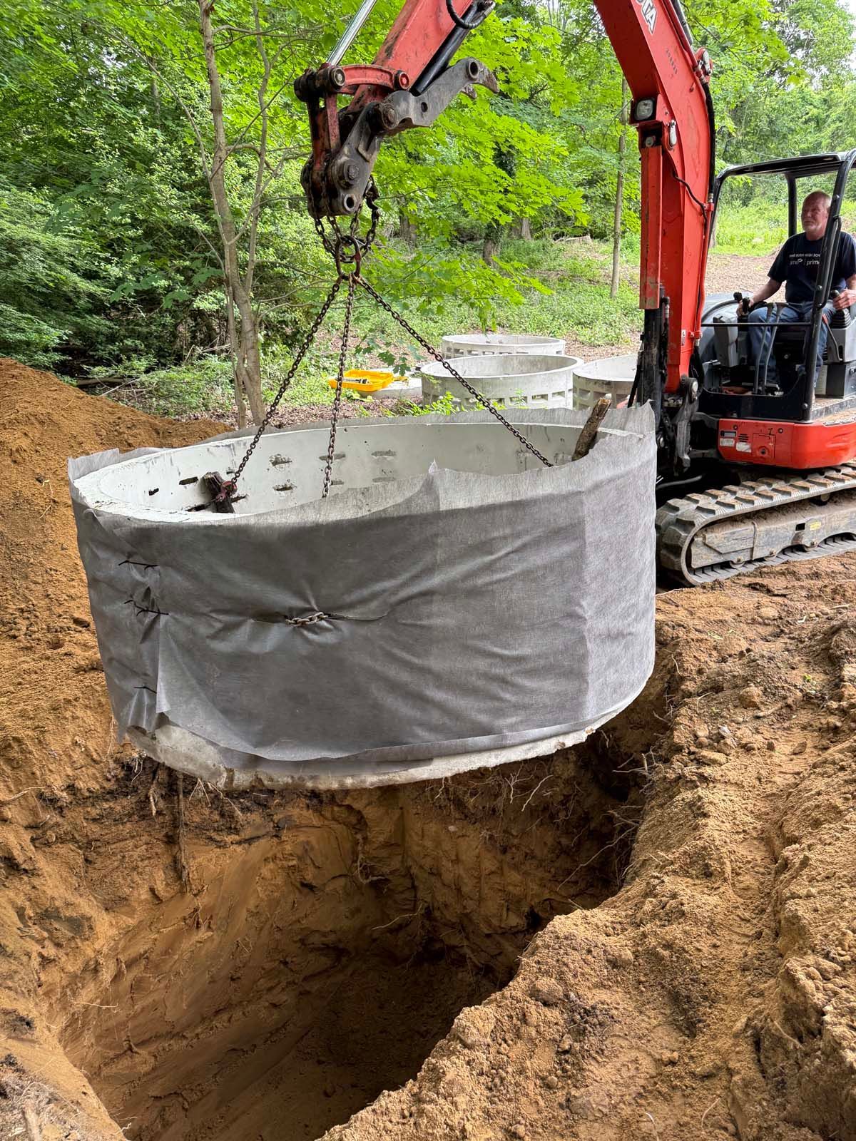 An excavator lowers a large concrete ring into a dug trench for construction.