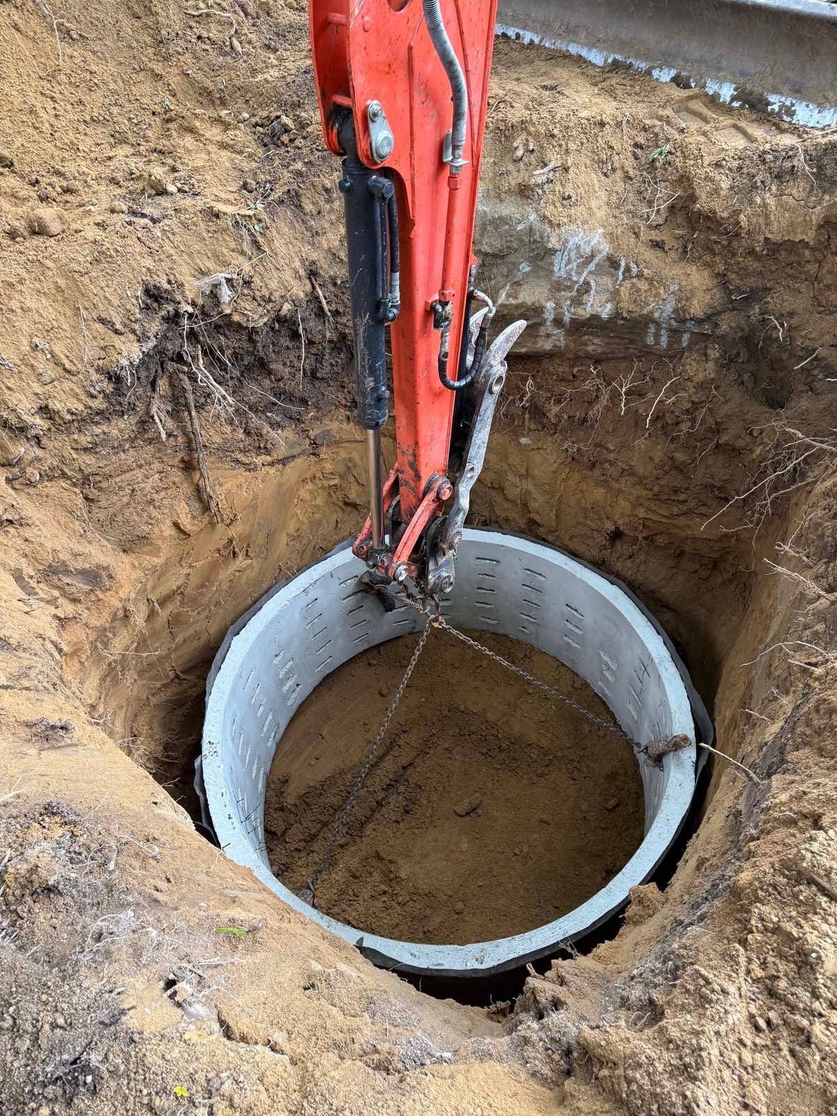 An excavator lowering a large concrete ring into a circular hole in the ground.