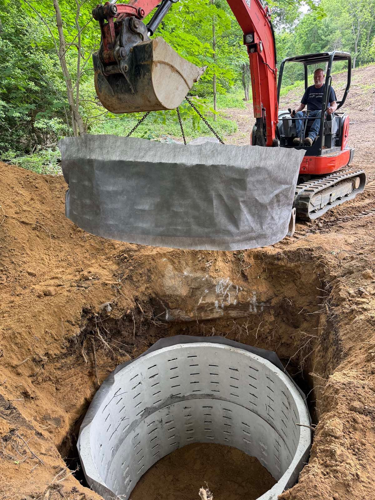An excavator lowers a concrete ring into a circular pit; a worker sits in the cab.
