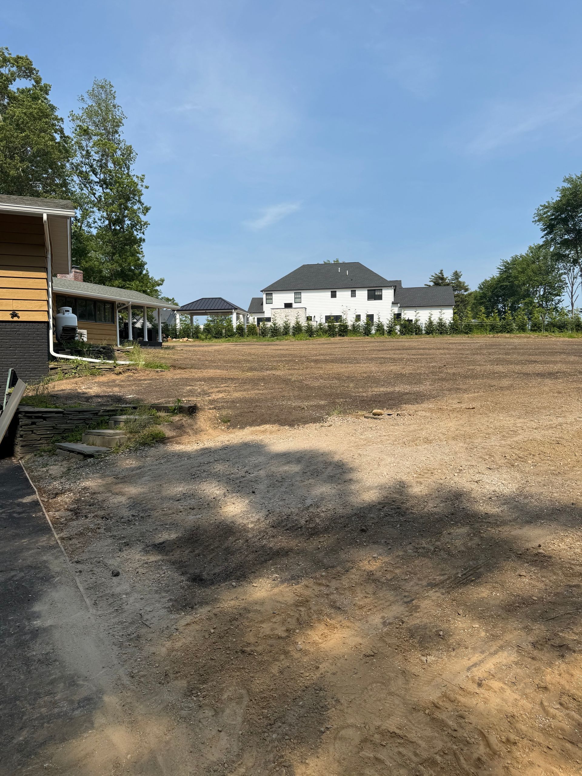A cleared lot with exposed soil, a partially built structure on the left, and a white house in the distance.