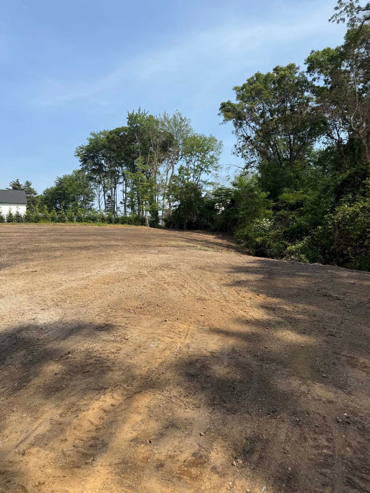Cleared dirt lot under a bright blue sky, with trees to the right and the left and a house visible on the left.