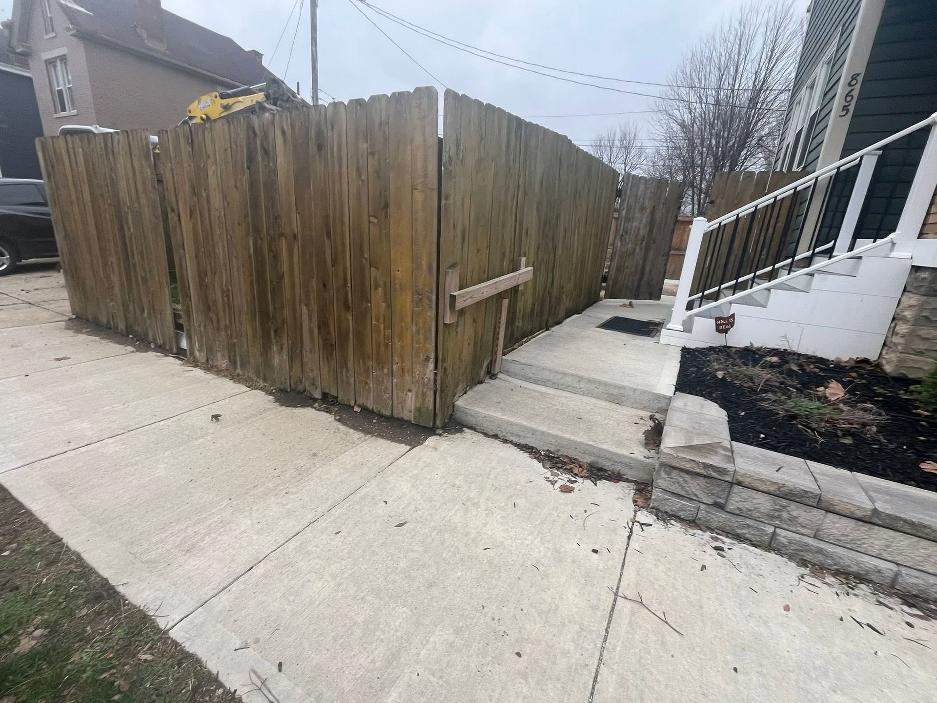 Wooden fence corner beside a concrete sidewalk and stairway with white railing.