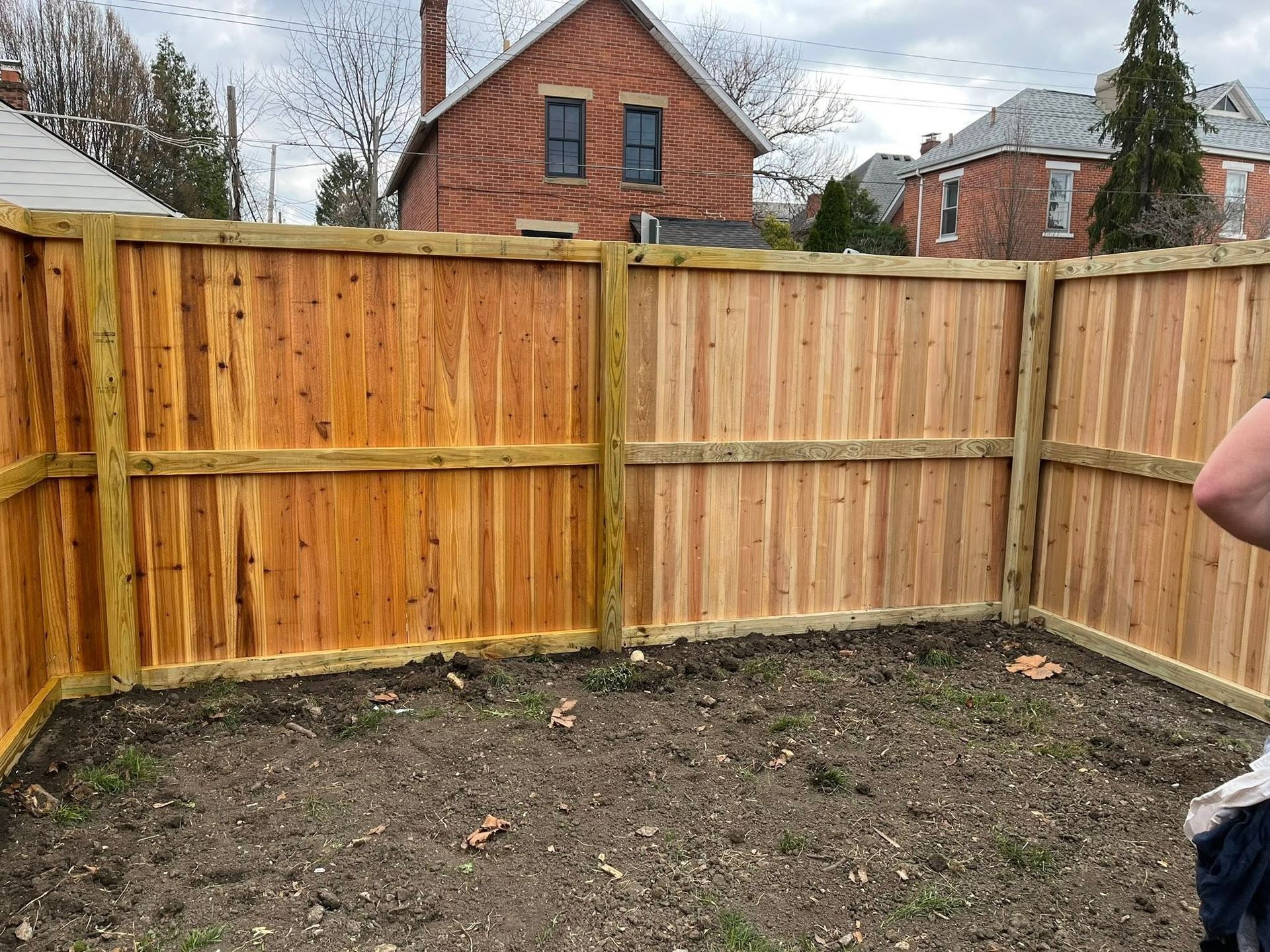 Small fenced backyard with bare soil and a wooden privacy fence, houses visible beyond it.