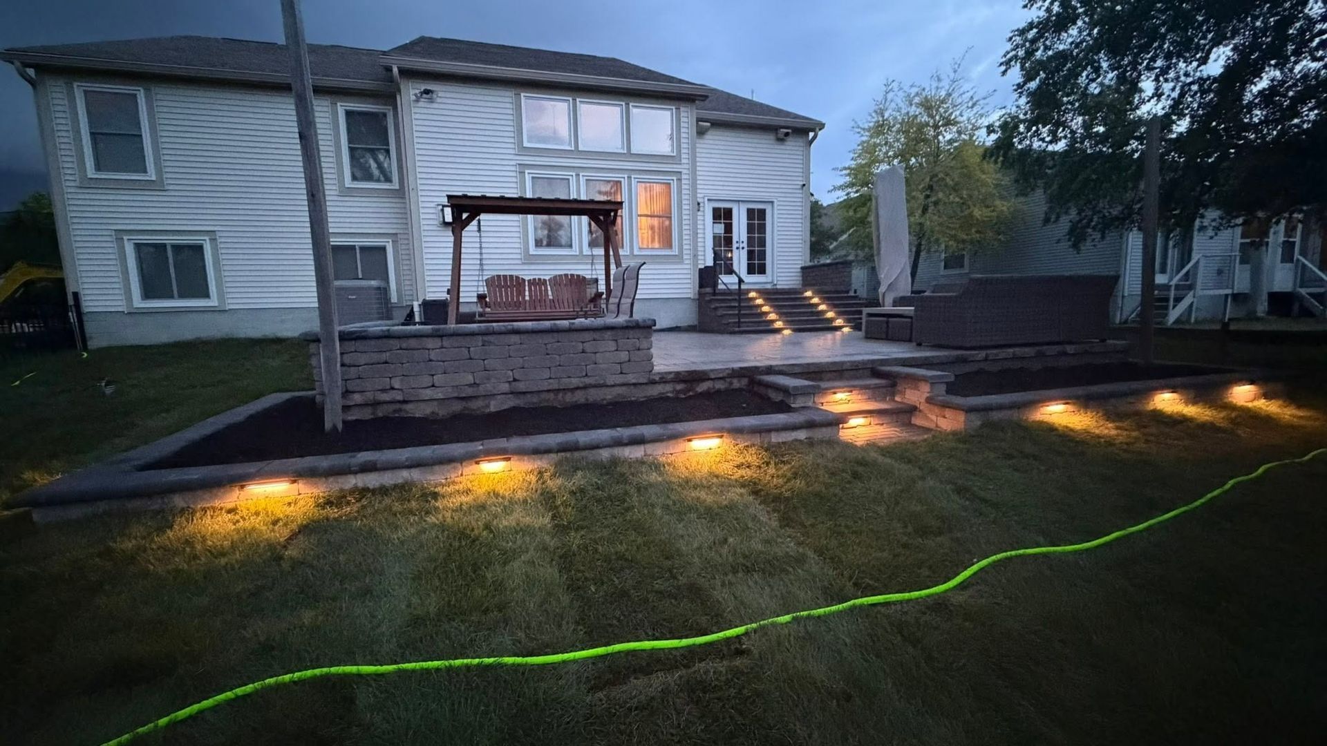 Backyard patio at dusk with warm ground lights, wet grass, and a two-story house in the background