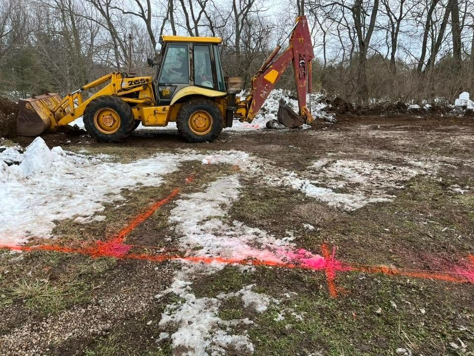 Yellow backhoe excavator at a muddy, snowy construction site with trees in the background