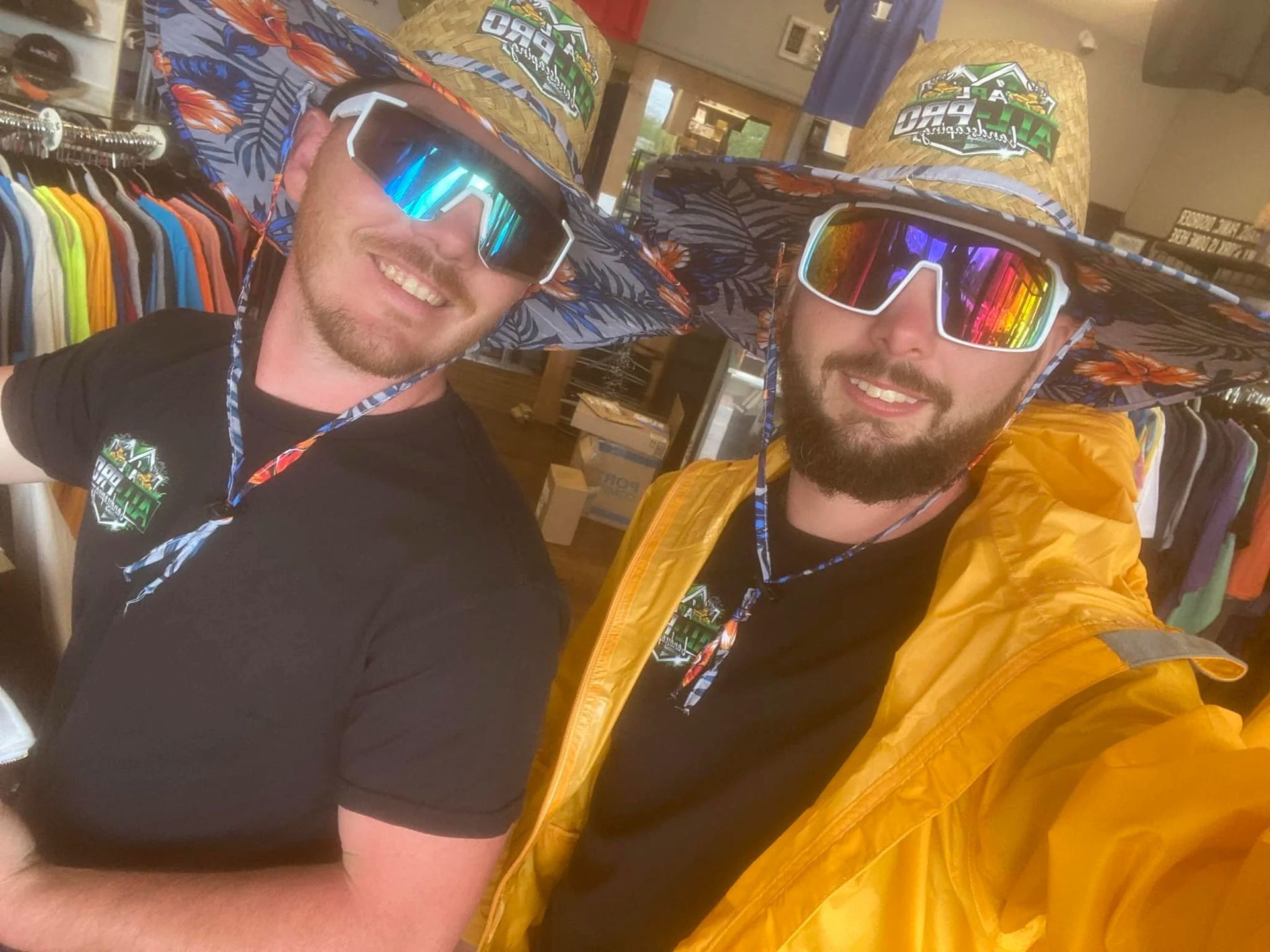 Two people smiling in a hat shop, wearing mirrored sunglasses and colorful hats; one wears a yellow jacket.