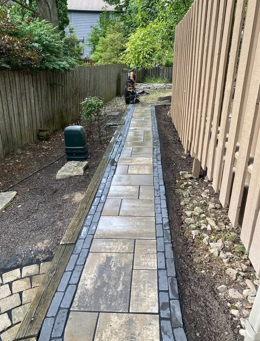 Narrow garden path with paving stones, bordered by wood fences and greenery, leading to a small green bin.