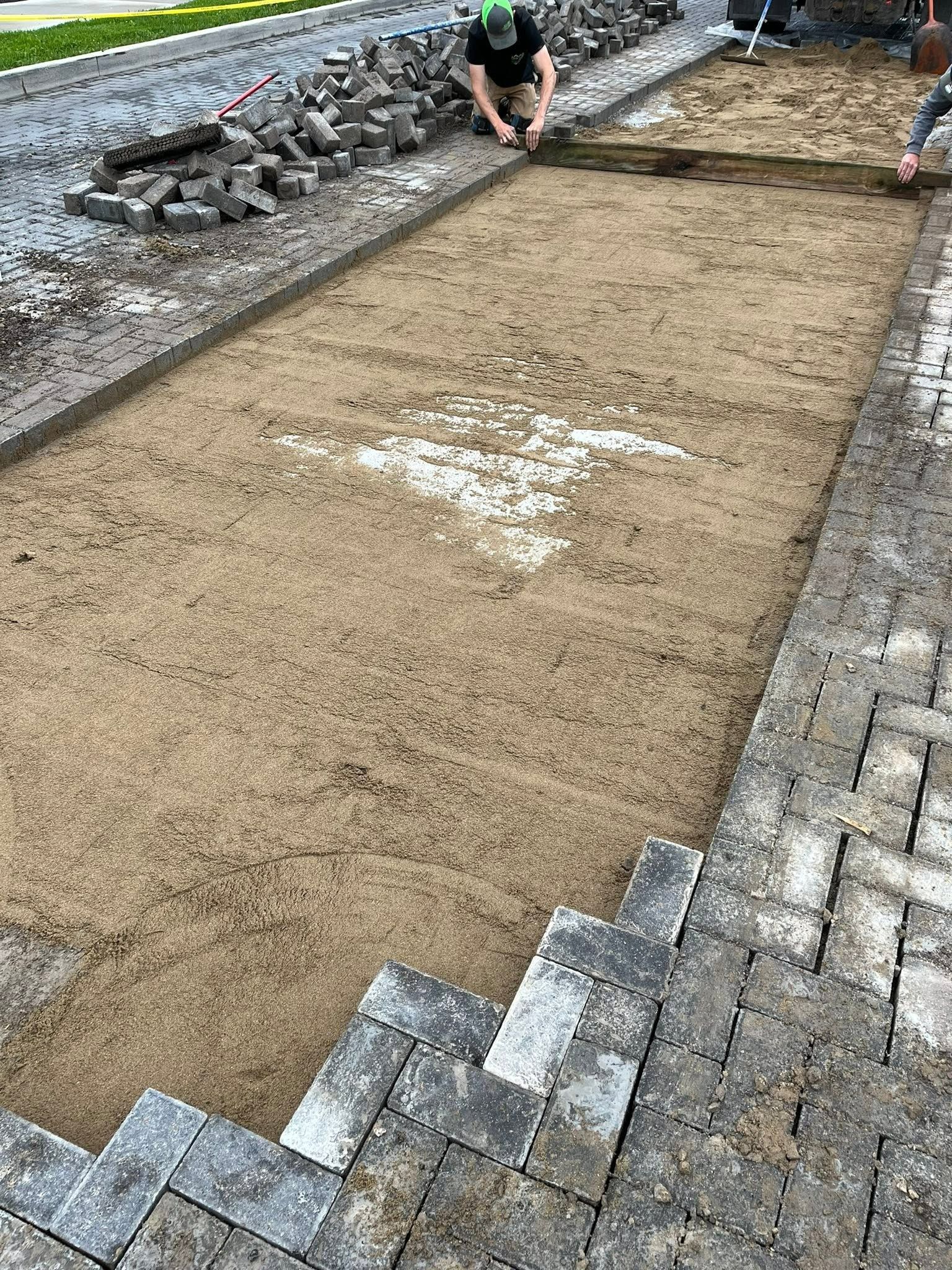 Rectangular sand area bordered by stone pavers, with workers smoothing the surface outdoors