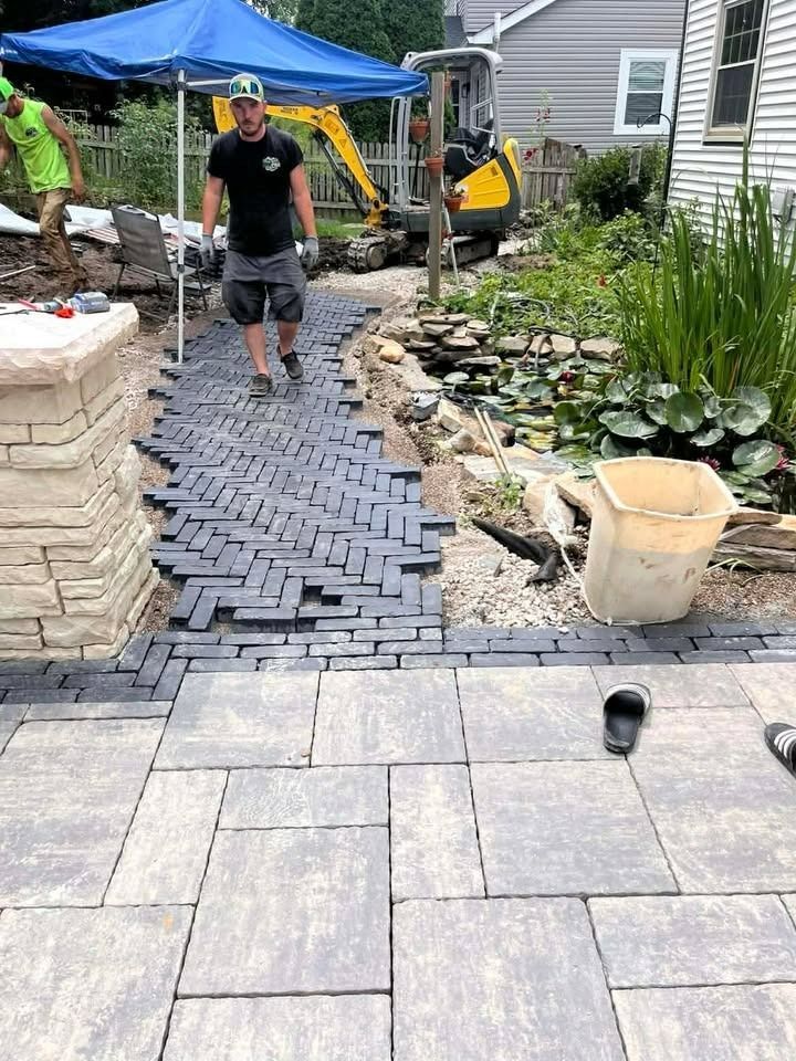 Worker laying dark pavers along a garden path beside a house and pondless water feature