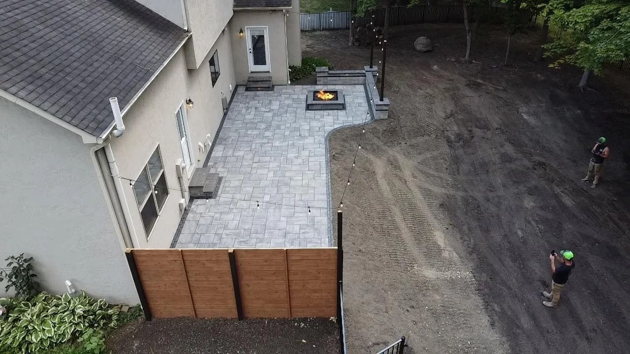 Aerial view of a backyard with stone patio, wooden fence, and two workers on a dirt area