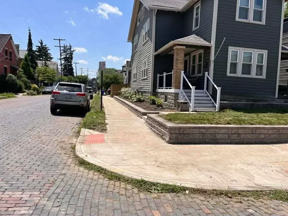Residential street with a parked car, cobblestone road, and a gray house with a front porch and ramp