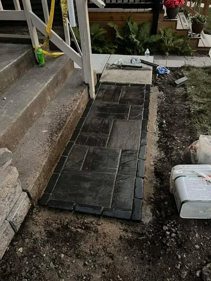Concrete paver walkway beside steps and porch, bordered by dirt and a white storage bin.