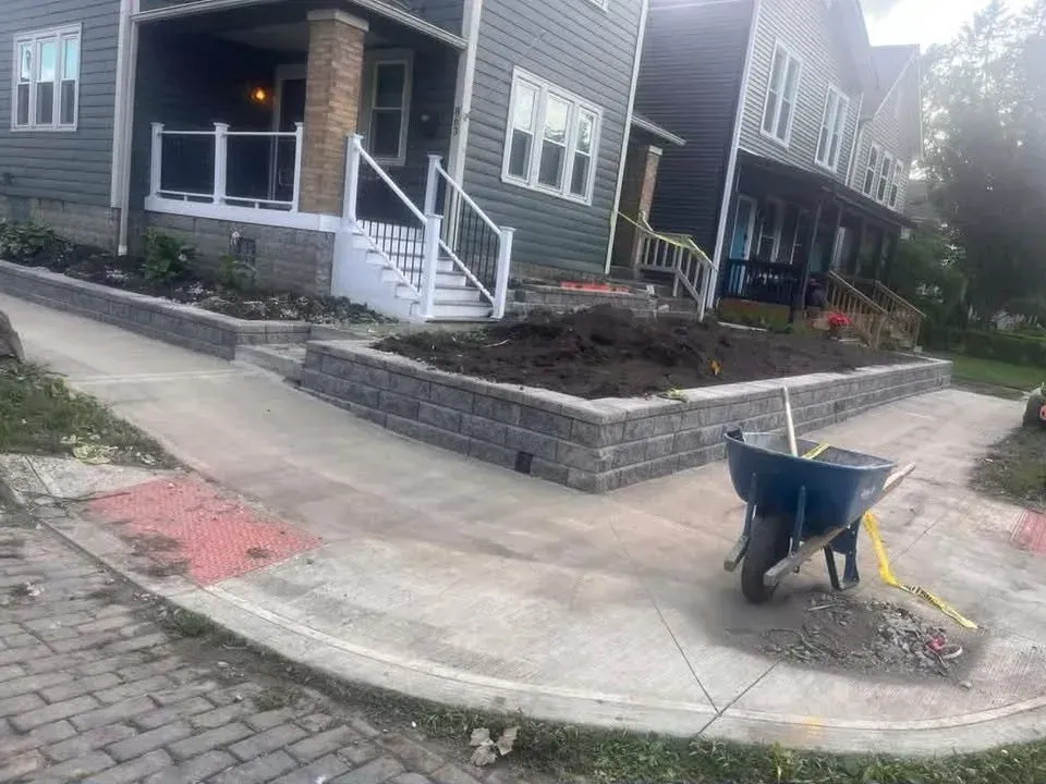 Row of townhouses with concrete sidewalk, retaining wall, and a wheelbarrow at the curb on a sunny day