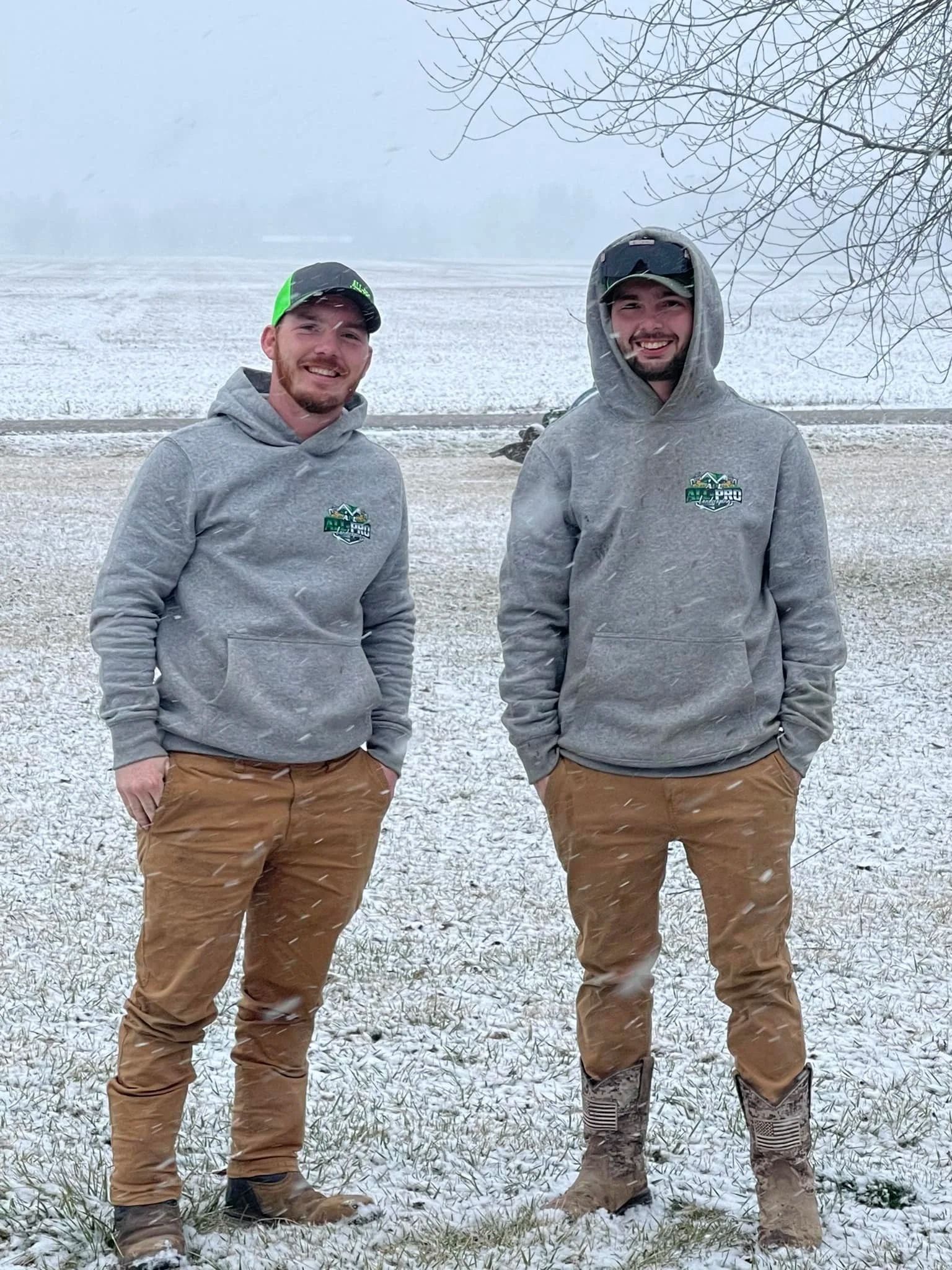 Two men in gray hoodies and brown pants stand in a snowy field with mountains in the background.