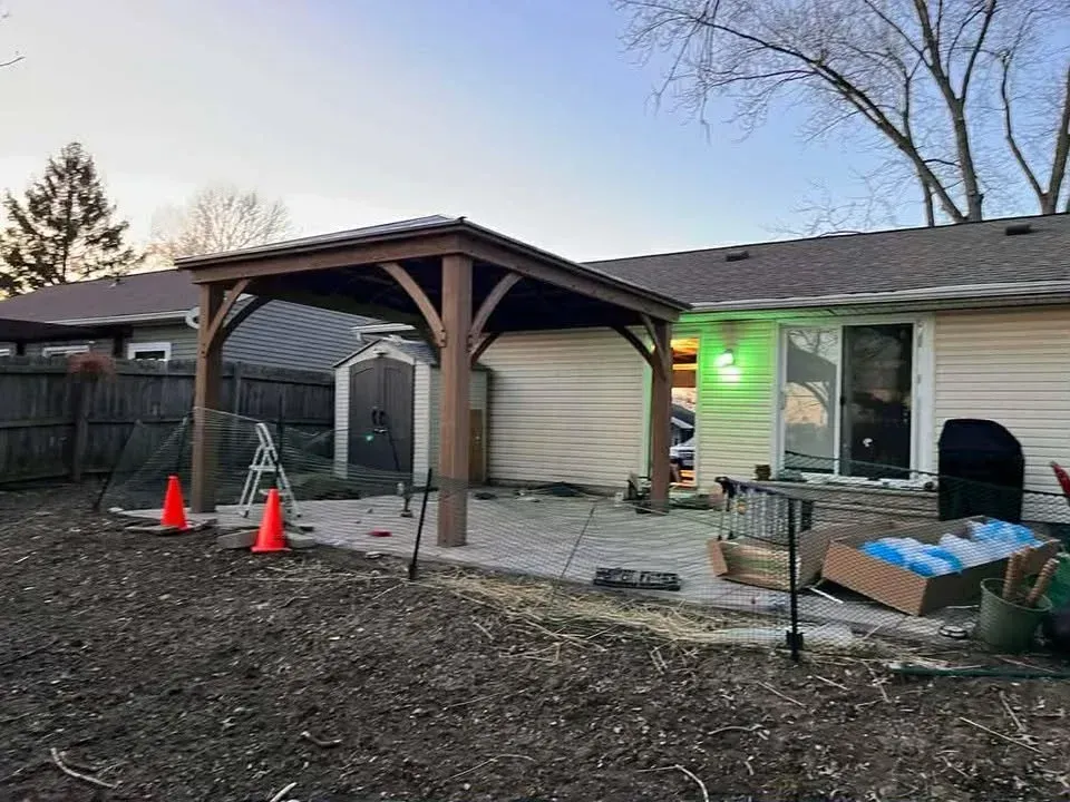 Backyard patio with wooden pergola, cones, green porch light, and a person working near a house.